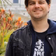 Lächelnder Mann in schwarzer Jacke und T-Shirt vor farbenfrohen Blumen und Gebäuden im Hintergrund.