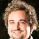 Ein lächelnder Mann mit lockigem, blondem Haar steht vor einem verschwommenen, grünen Hintergrund.