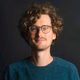 Ein lächelnder Mann mit lockigem, blondem Haar und Brille trägt ein blaues Sweatshirt vor einem dunklen Hintergrund.