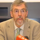 Ein Mann mit Brille und grauem Bart spricht bei einer Konferenz, gekleidet in einem hellen Anzug.