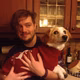 Ein junger Mann hält einen Hund in einem roten Pullover. Der Mann lächelt, die Umgebung ist eine Küche mit Holzschränken.