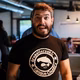 Mann mit Bart, der ein schwarzes T-Shirt mit einem Grafiklogo trägt, zeigt Freude in einem Innenraum mit Menschen im Hintergrund.