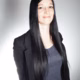 Frau mit langen, schwarzen Haaren und einem schwarzen Blazer, lächelnd vor einem hellen, neutralen Hintergrund.