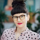 Frau mit Brille und schwarzem Haarknoten, trägt ein gepunktetes Hemd und lächelt. Im Hintergrund sind Pflanzen sichtbar.