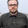 Ein Mann mit Brille und Bart trägt ein schwarzes Hemd und schaut direkt in die Kamera. Der Hintergrund ist grau und unscharf.