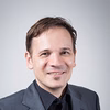 Ein Mann mit kurzer, brauner Frisur, trägt einen dunklen Anzug und lächelt freundlich in die Kamera. Der Hintergrund ist hell und neutral.