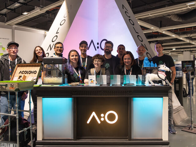 A group of people standing behind a black booth with illuminated white panels, displaying "A:O" logo, at an exhibition with a white triangular backdrop.