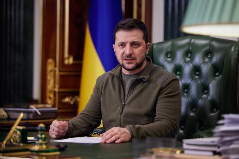 Ukrainian president sitting at a desk with a serious expression, wearing a green jacket, with a Ukrainian flag behind him.