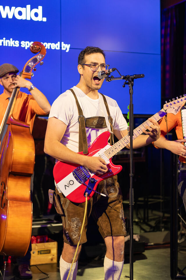 A musician wearing traditional Austrian attire sings into a microphone while playing a bright red guitar with ACOLONO logo.