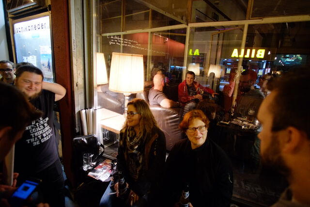 A group of people gathered inside a cozy bar with warm lighting, some sitting on couches and others standing, enjoying drinks and conversation at night.