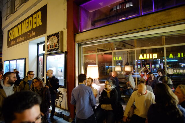 Crowd of people gathering outside a venue with a sign reading "Schraneder Kino Bühne Cafe" at night; some are standing and chatting, others waiting or entering.