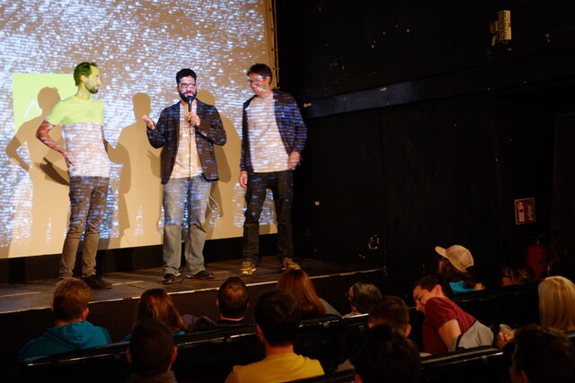 Three men standing on a stage, one speaking into a microphone, with a large audience seated in front, and a projection screen behind them.