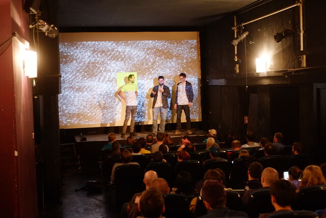 Three men stand on a stage in front of a large screen, one holding a microphone and speaking, while the audience watches attentively.
