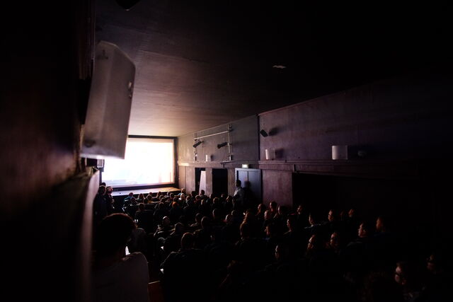 People sitting in a dark theater watching a large screen, with some standing or entering the room, and natural light coming from a window on the left.