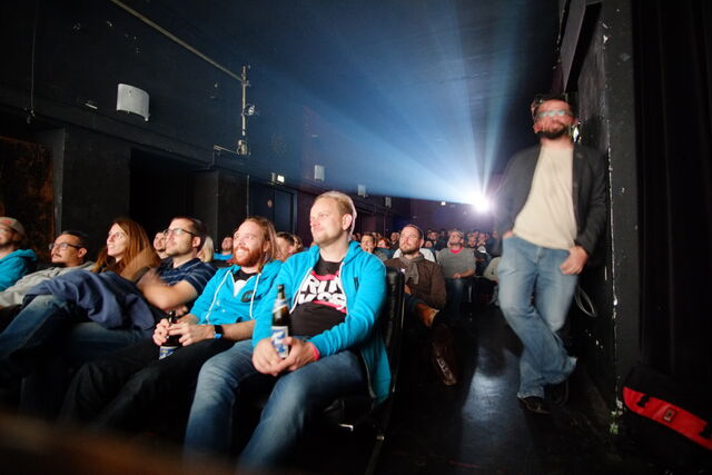 People sitting in an audience watching a screen, some smiling and holding drinks, with a person standing near the wall on the right.