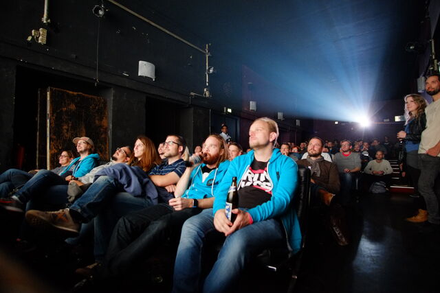 A group of people sitting in a dark theater, watching a performance or screening, with focused and engaged expressions; some holding drinks.