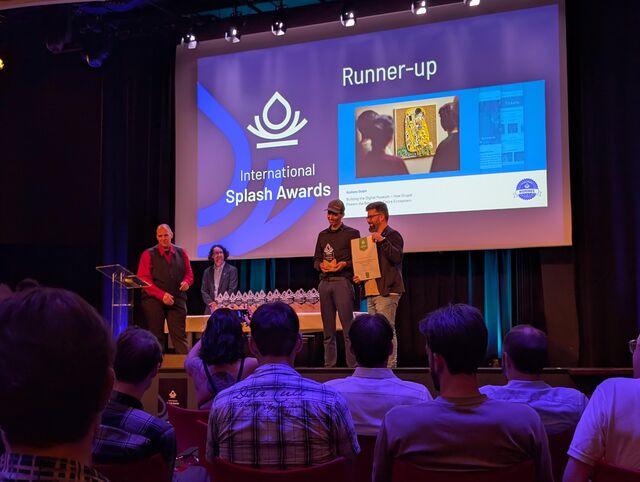 Zwei Personen stehen auf der Bühne und halten einen Preis, während die Zuschauer im Vordergrund sitzen. Im Hintergrund werden die Splash Awards präsentiert.