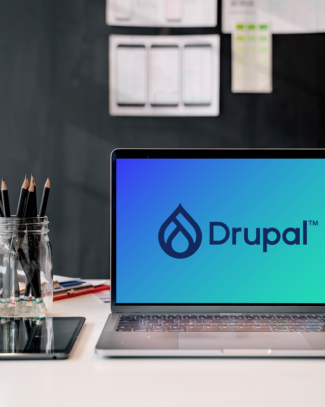 Ein Laptop mit dem Drupal-Logo auf dem Bildschirm, umgeben von einem Glas mit Bleistiften und einem Tablet auf einem Schreibtisch.