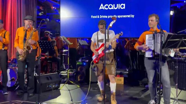 A band performing on stage with musicians playing saxophone, guitar, and accordion, in front of a large screen displaying "Acquia" and event details.