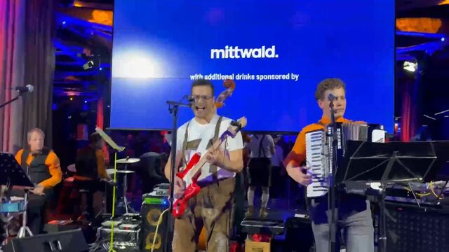A band performs on stage with a large blue screen behind them displaying "mittwald" and a message about sponsored drinks, surrounded by colorful lighting.