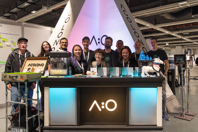 A group of people standing behind a black booth with illuminated white panels, displaying "A:O" logo, at an exhibition with a white triangular backdrop.