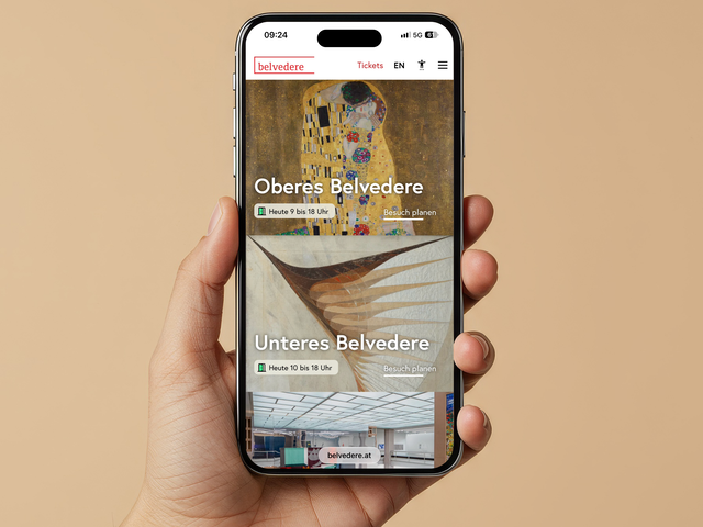 Eine Hand hält ein Smartphone, das die Website des Belvedere mit Kunstwerken auf einem neutralen Hintergrund zeigt.