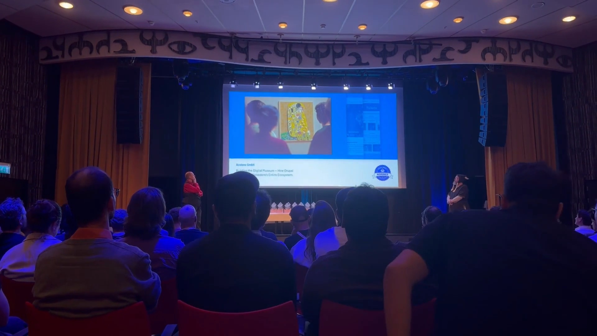 Eine Präsentation in einem Raum mit Publikum, auf einer Leinwand wird eine Grafik angezeigt, während Sprecher aktiv sind.